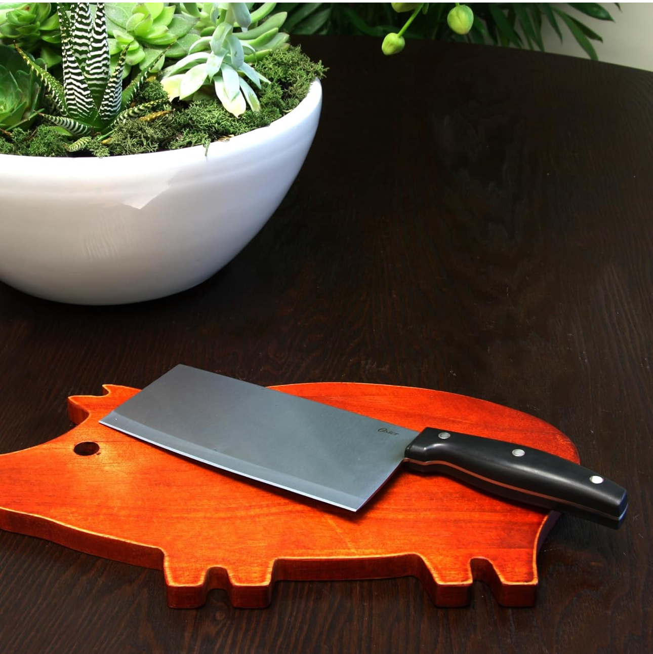 Pig-shaped cutting board with a knife on a wooden surface next to a white bowl with plants.